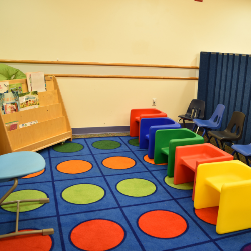 An empty ABA group therapy room with colorful, kid-sized chairs, a shelf of books, a chair for the therapist, and a blue rug with large colorful dots.