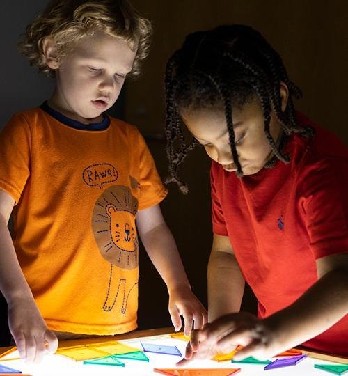 two young boys play with colorful shape tiles on a lighted table