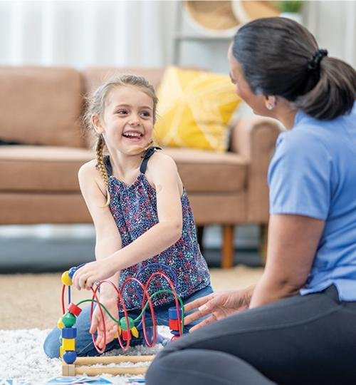 A young girls plays with toys on the floor next to her female therapist