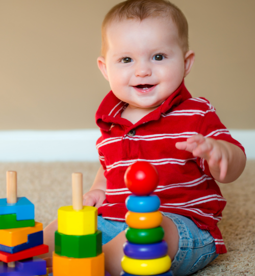 A smiling baby boy plays with colorful blocks while sitting on the floor