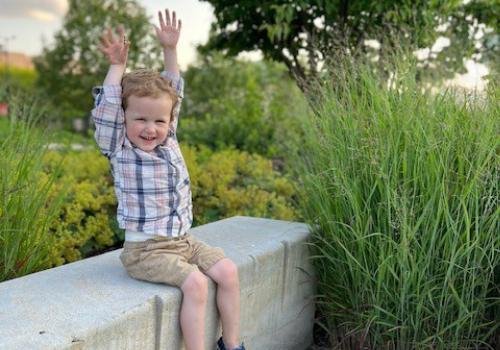 A happy, young boy in a blue plaid shirt sitting outside on a wall with his hands up in the hair