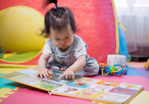 A small baby with black hair looking at a book on a colorful carpet